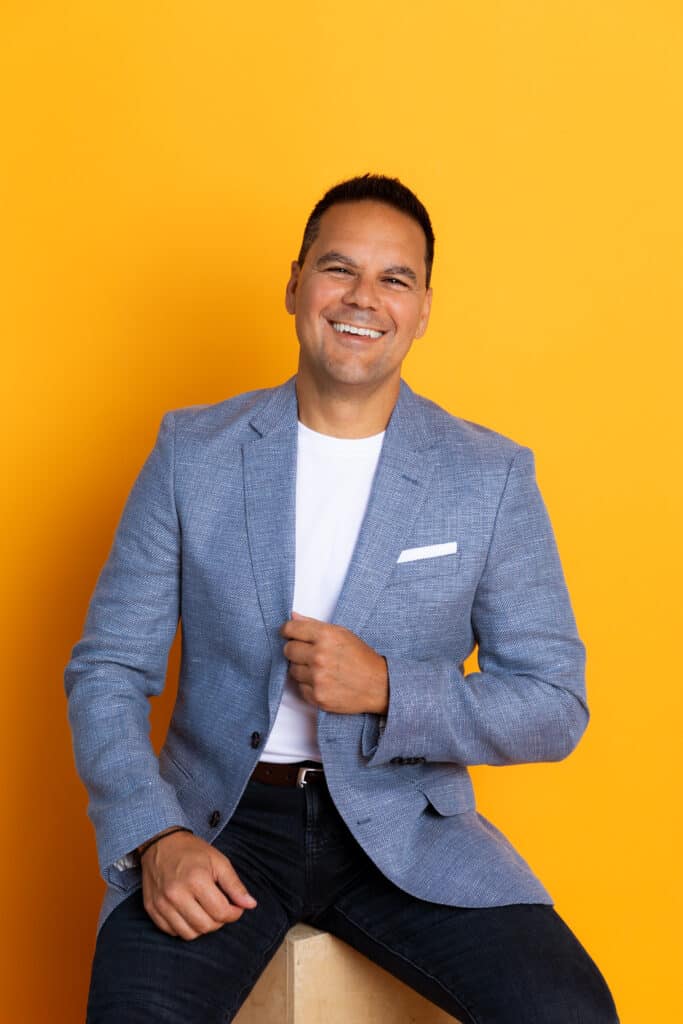 Joe Stella in a light blue blazer and white t-shirt sitting against a vibrant yellow background.