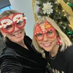 Two women wearing festive holiday glasses, one with a snowman design and the other with Christmas trees, smiling in front of a decorated Christmas tree with gold ribbon and ornaments.