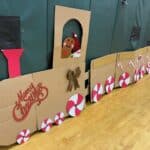 A handmade cardboard Christmas train decorated with red and white peppermint wheels, candy canes, a gingerbread figure wearing a Santa hat, and a festive bow, with "Merry Christmas" written on the front.