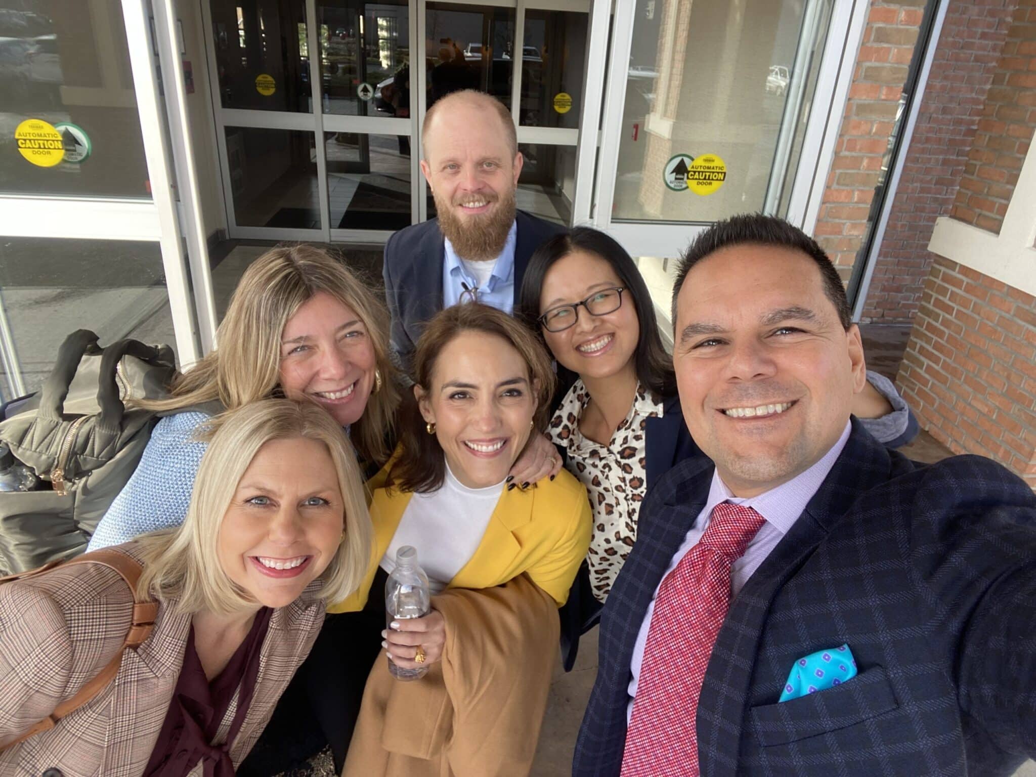 A group of six professionally dressed individuals smiling as they take a selfie outside a building entrance, with luggage visible in the background.