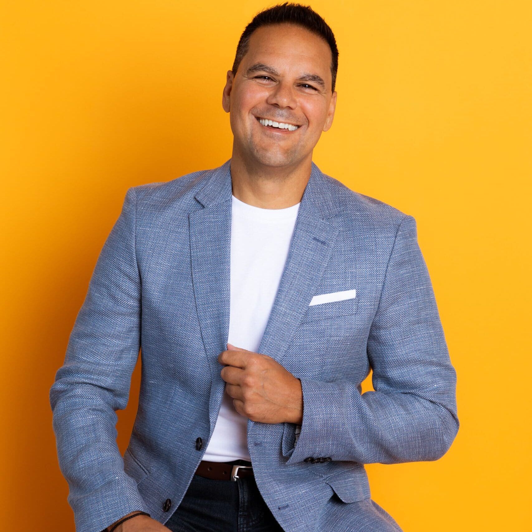 Joe Stella in a light blue blazer and white t-shirt sitting against a vibrant yellow background.