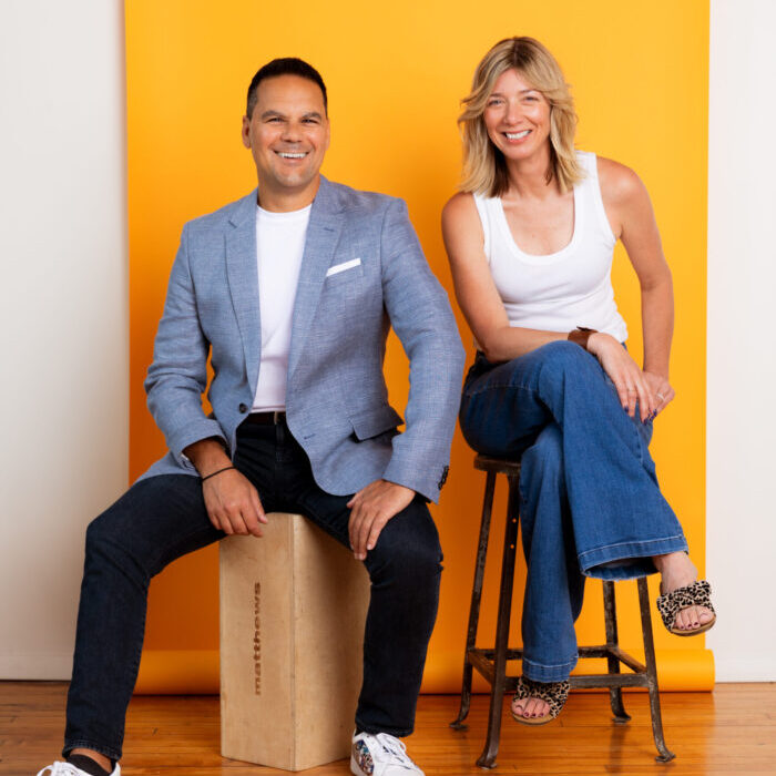 Studio photo of Joe Stella and Rene Ryan smiling on stools against a yellow backdrop.