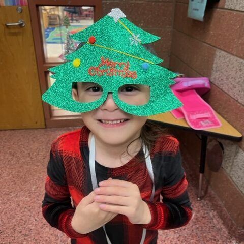 A young smiles while wearing green Christmas tree glasses reading "Merry Christmas."
